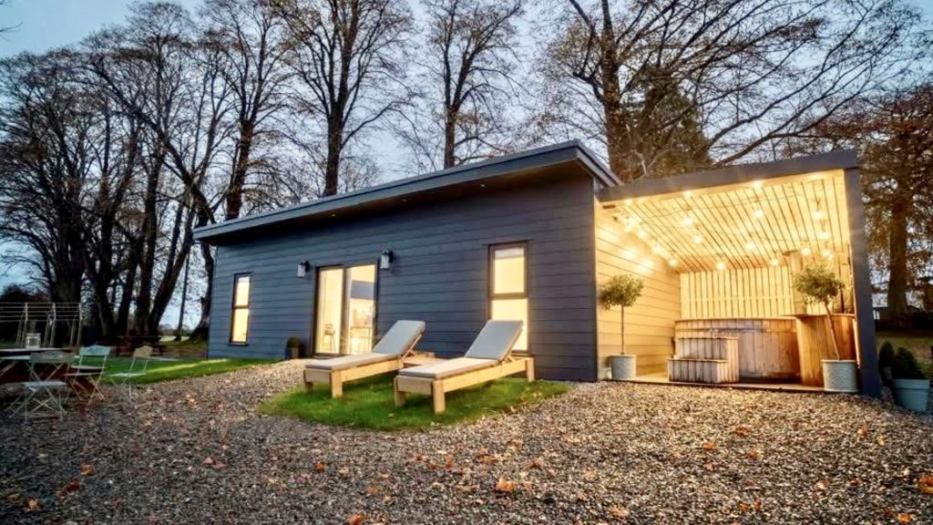 Image of a lodge, dimly lit at dusk with a covered hot tub, at Brecart Lodge.